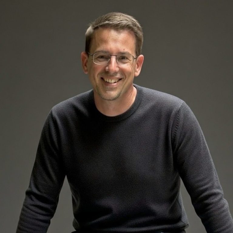 Lächelnder Mann in einem schwarzen Pullover vor neutralem Hintergrund.